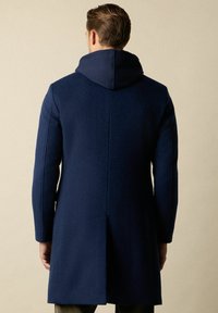 Granatowy wełniany płaszcz z kapturem, o prostym kroju, bezszwowych plecach i subtelnym teksturowanym wykończeniu. Styl wyposażony w boczne rozcięcia dla swobody ruchów.