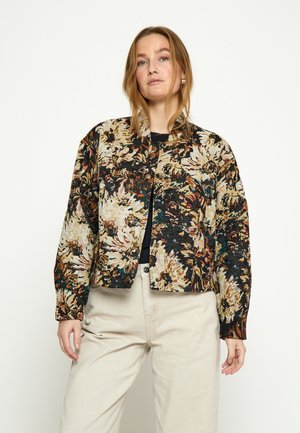 Mujer vistiendo una chaqueta corta con patrón floral en tonos oscuros, beige y marrón sobre una camiseta negra y pantalones beige claros, de pie contra un fondo liso.