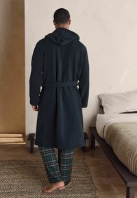 Temne zelený kapucňový plášť vyrobený z jemnej látky, s dlhými rukávmi a zaväzovaním v páse. V kombinácii s nohavicami s károvaným vzorom.