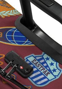 Valigia a rotelle con tessuto color bordeaux, decorata con toppe a tema viaggio, impugnatura telescopica e un lucchetto nero per la sicurezza.
