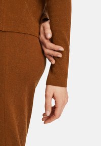 Braun gestricktes Outfit mit einem subtilen Muster, das eine strukturierte Oberfläche aufweist. Eine Hand ruht sanft auf dem Arm und betont die weiche, figurbetonte Silhouette.