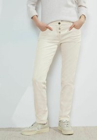 Beżowe bawełniane spodnie o prostych nogawkach, z czterema kieszeniami i zapięciem na guziki. W połączeniu z jasnymi trampkami, na jednolitym tle.