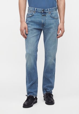 Uomo che indossa jeans azzurri a taglio dritto con camicia blu infilata e scarpe di pelle nere su sfondo bianco.