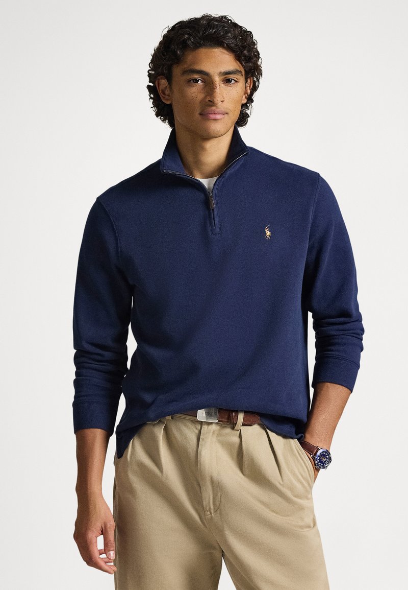 Male model wearing navy blue quarter-zip sweater and beige pants, with a brown belt and blue wristwatch, standing against a white background.
