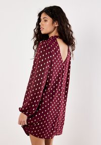 Donna con lunghi capelli ricci che indossa un abito bordeaux con pois dorati e una scollatura a V profonda, in piedi su uno sfondo bianco uniforme.