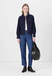Casaco de denim escuro com fecho de botão, dois bolsos no peito; combinado com jeans azul médio, botas pretas de salto e uma mala preta.