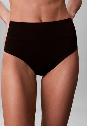 Kobieta w czarnych, bezszwowych majtkach z wysokim stanem, dłonie spoczywają na biodrach, pokazana od środka tułowia do górnej części ud.