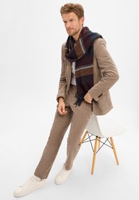 Beige corduroy suit with a white shirt, paired with a patterned dark scarf. White sneakers and sitting on a modern wooden chair.