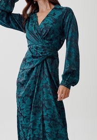 Mörk teal satin klänning med lång ärm och blommönster, med ett omslagdesign och rynkad midja för en figurnära siluett.