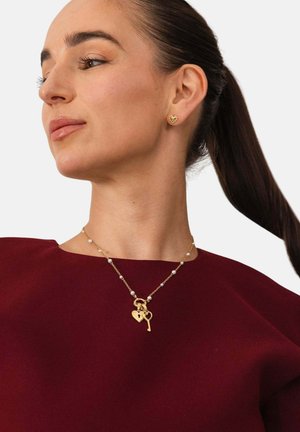 Woman with dark hair in a ponytail wearing a burgundy top, gold heart-shaped earrings, and a gold necklace with heart and key charms.