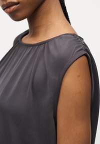 Gros plan d'une personne portant un haut sans manches gris foncé avec un tissu froncé au niveau de l'encolure et des épaules.