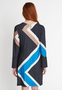 Langärmliges Kleid in Marineblau, mit geometrischen Mustern in Creme, Braun und Blau. Schlüssel-Ausschnitt hinten; weicher, glatter Stoff.