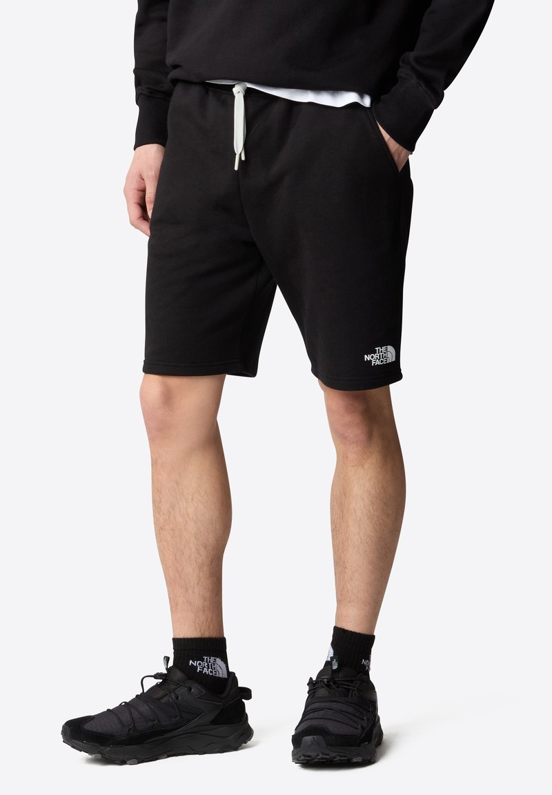 Pantalones cortos de algodón negros con cintura elástica y cordón, con bolsillos laterales y un logo blanco en el dobladillo izquierdo, combinados con zapatos negros.