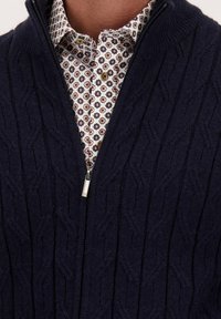 Man in een marineblauwe kabelgebreide zip-up trui over een wit shirt met een veelkleurig cirkelpatroon.