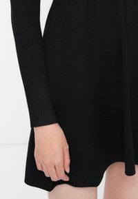 Gros plan sur la main d'une personne reposant à côté d'une robe noire en maille côtelée à manches longues avec une jupe évasée.