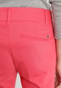 Vaaleanpunaiset housut, joissa on sileä pinta, sisältävät takataskun vetoketjulla ja pienen koristeellisen logon. Vyötäröllä on vyölenkit.