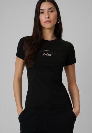 Kobieta o długich ciemnych włosach, ubrana w czarny dopasowany t-shirt z napisami „komfort w każdym ruchu” i „poczuj się przytulnie”, stojąca z rękami w kieszeniach.