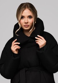 Černá péřová bunda s vysokým límcem, širokými rukávy a opaskem v pase. Tkanina vypadá hladce s matnou strukturou.