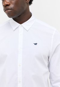 Biała koszula z guzikami i klasycznym kołnierzykiem, z małym niebieskim haftowanym logo konia na piersi oraz gładką bawełnianą teksturą.