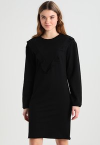 Schwarzes langärmeliges Kleid mit einem V-Ausschnitt und Rüschen sowie einer figurschmeichelnden Silhouette, gefertigt aus weichem, angenehmem Stoff.