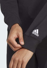 Černá mikina se žebrovanými manžetami, na rukávu se nachází bílé logo adidas. Tkanina působí hladce a jemně na pokožce.