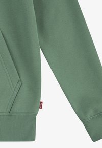 Manche et poche de sweat-shirt vert avec poignets et ourlet côtelés, avec une petite étiquette rouge de la marque Levi's.