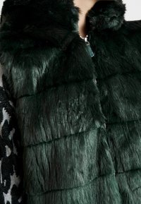 Dunkelgrüne Kunstpelzweste mit horizontalen Streifen, hohem Kragen und silbernem Zipper-Detail. Weiche Textur mit geschichtetem Design.