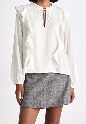 Mujer que lleva una blusa blanca de manga larga con volantes y cuello con lazo, combinada con una minifalda gris de cuadros.