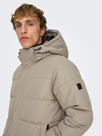 Beige puffer jacket featuring a tall hood, zippered pockets, and a quilted pattern. Soft texture with black accents on sleeves and hood.
