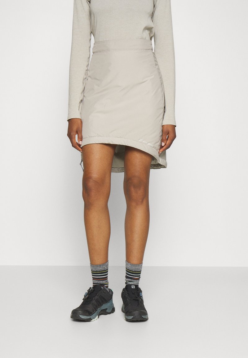 Jupe beige mi-longue avec des détails de resserrement sur le côté, portée sur des chaussures de sport noires et des chaussettes rayées. Texture lisse et design moderne.