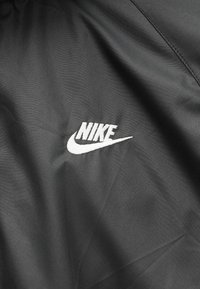 Μαύρο ύφασμα με λευκό κεντημένο λογότυπο Nike που περιλαμβάνει το όνομα της μάρκας και το σύμβολο swoosh στην κεντρική περιοχή.