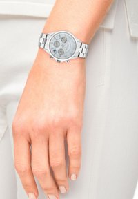 Zilveren chronograaf horloge met een ronde wijzerplaat, metalen band, meerdere wijzerplaten en een gestructureerde achtergrond. G gedragen op een lichtgekleurde hand.