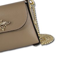 Beige leren schoudertas met gouden kettingriem, met een decoratief bijembleem op de flap en zwarte afwerking rond de randen.
