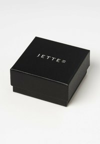 Svart smyckeskrin med en texturerad yta, med logotypen "JETTE" präglad i silver på toppen. Fyrkantig form med lock som har gångjärn.