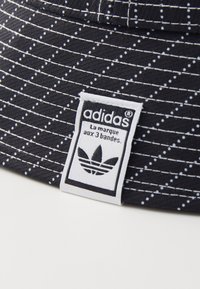 Svart bucket-hatt med en texturerad tyg med vita diagonala ränder och prickar. Visar en Adidas-logotyp på framsidan.