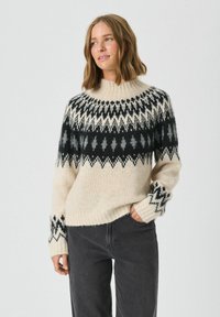 Sweter w kolorze kremowym, dzianinowy, z wysokim kołnierzem, czarno-szarymi wzorami geometrycznymi oraz ściągaczami przy mankietach. W połączeniu z ciemnoszarymi spodniami.