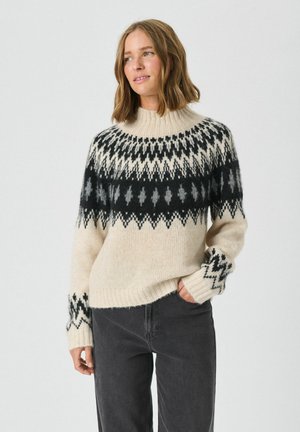 Cremfarbener Strickpullover mit hohem Kragen, schwarzen und grauen geometrischen Mustern sowie gerippten Bündchen. Kombiniert mit dunkelgrauen Hosen.