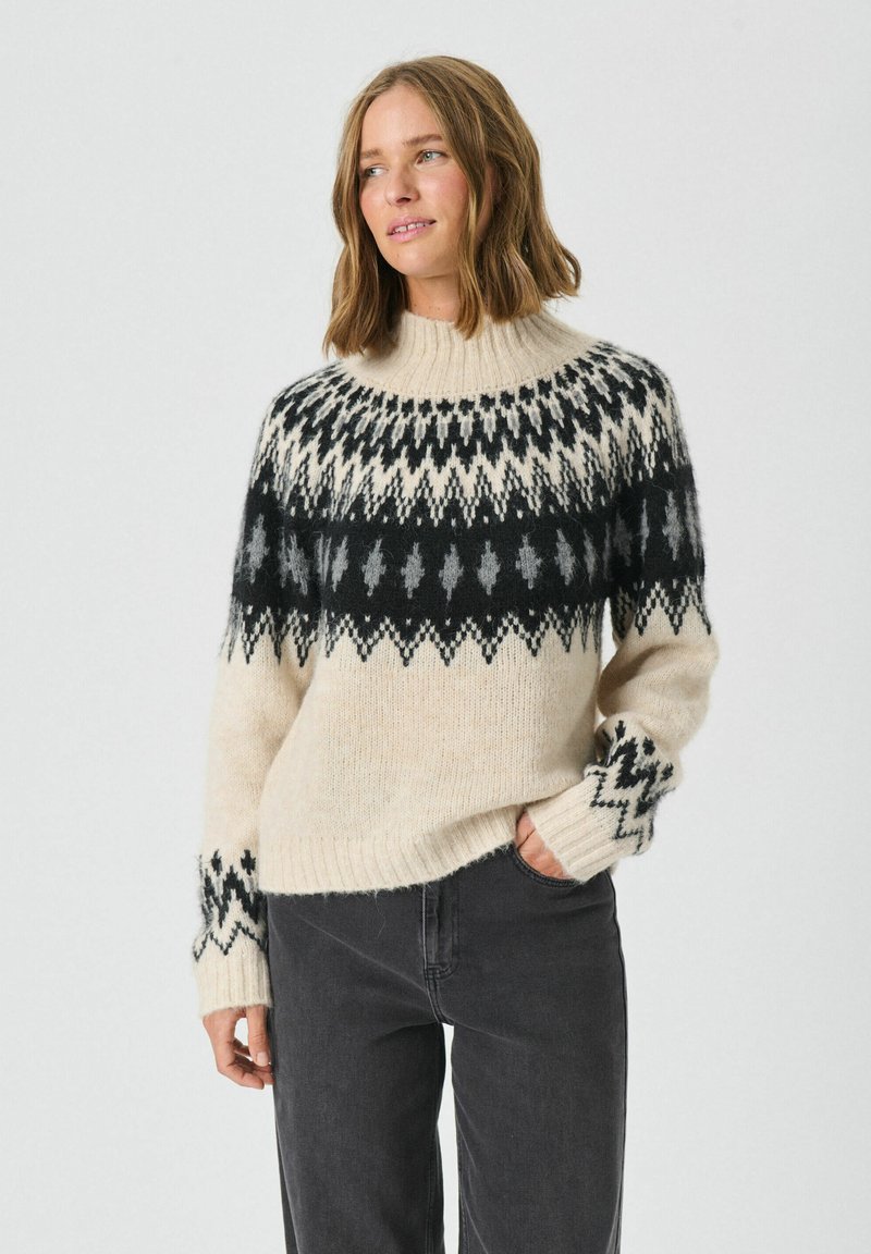 Cremfarbener Strickpullover mit hohem Kragen, schwarzen und grauen geometrischen Mustern sowie gerippten Bündchen. Kombiniert mit dunkelgrauen Hosen.