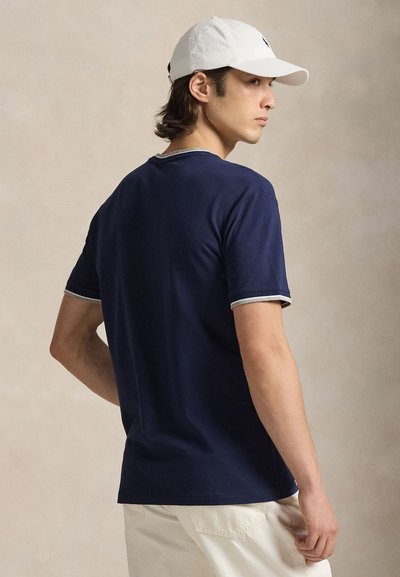 Mladý muž nosiaci tmavomodré tričko so sivým lemom, biele nohavice a bielu šiltovku, stojaci pred jednoliatym béžovým pozadím.