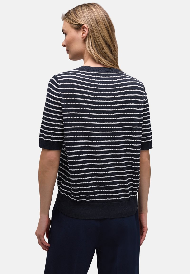 Femme aux cheveux blonds portant un haut à manches courtes rayé bleu marine et blanc et un pantalon foncé, vue de dos sur un fond uni.