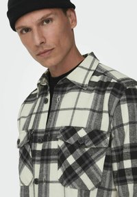 Camisa de franela a cuadros en blanco y negro con dos bolsillos en el pecho, cierre de botones y textura de tela contrastante. El modelo lleva un gorro negro.