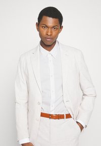 Helle beige Anzugjacke aus strukturiertem Stoff, kombiniert mit einem weißen Hemd und einem braunen Leder-Gürtel. Die Passform ist maßgeschneidert, mit zwei vorderen Knöpfen.