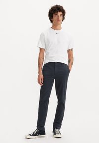 Hvid bomulds-t-shirt, navy bomuldsbukser og sorte canvas sneakers med hvide såler. Outfitet har en simpel pasform og et afslappet udseende.
