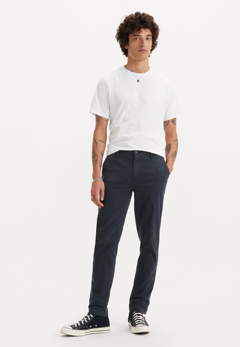 Hvid bomulds-t-shirt, navy bomuldsbukser og sorte canvas sneakers med hvide såler. Outfitet har en simpel pasform og et afslappet udseende.
