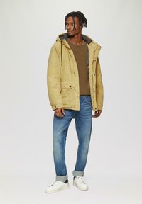 Beige hooded jack met trekkoorden, gelaagd over een gestructureerde bruine trui. Gecombineerd met lichtblauwe spijkerbroek en witte sneakers.
