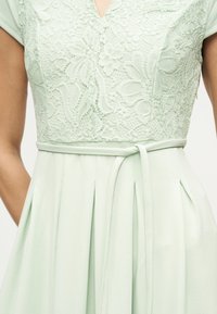 Φόρεμα ανοιχτού πράσινου χρώματος με μπούστο από λουλουδάτη δαντέλα, κοντά μανίκια, V-λαιμόκοψη και λεπτή ζώνη δεμένη στη μέση.