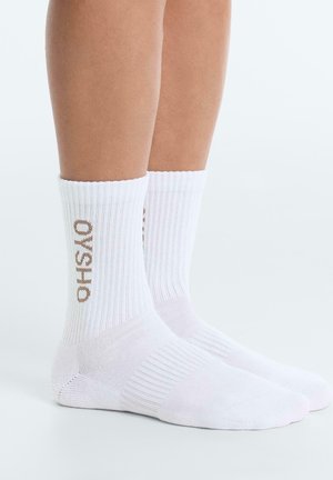 Picioare purtând șosete albe drepte cu dungi și text maro „OYSHO” pe părți, stând pe o suprafață albă.