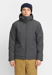 Chaqueta gris aislante con cuello alto, cremallera completa y capucha. Presenta puños elásticos y bolsillos laterales. Lleva puesta una boina de punto azul marino.