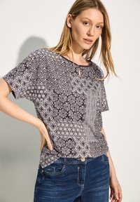 Sorte og hvide mønstrede kortermede top med geometriske designs og nøglehulshals, parret med blå jeans. Glat bomuldsstof.