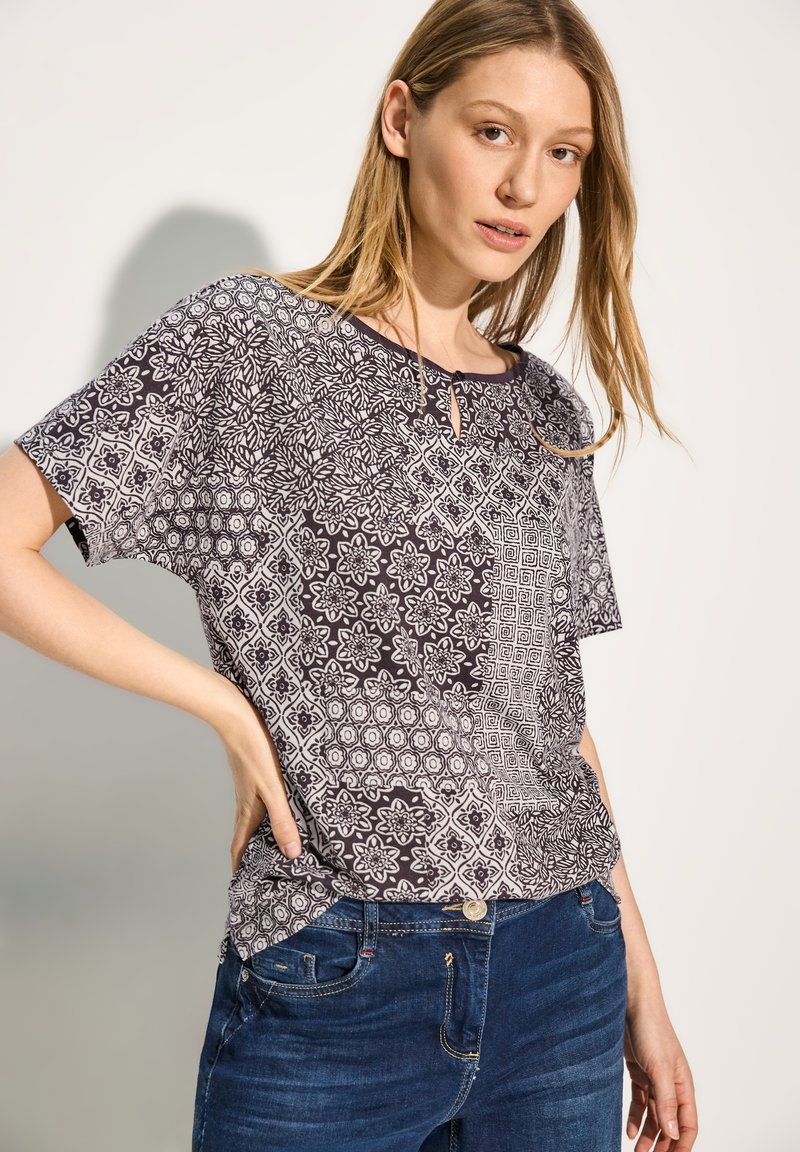Sorte og hvide mønstrede kortermede top med geometriske designs og nøglehulshals, parret med blå jeans. Glat bomuldsstof.